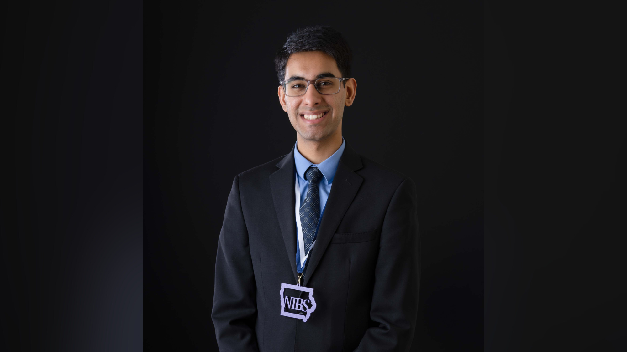 Agam Sharma smiles by a black background wearing a NIBS medal