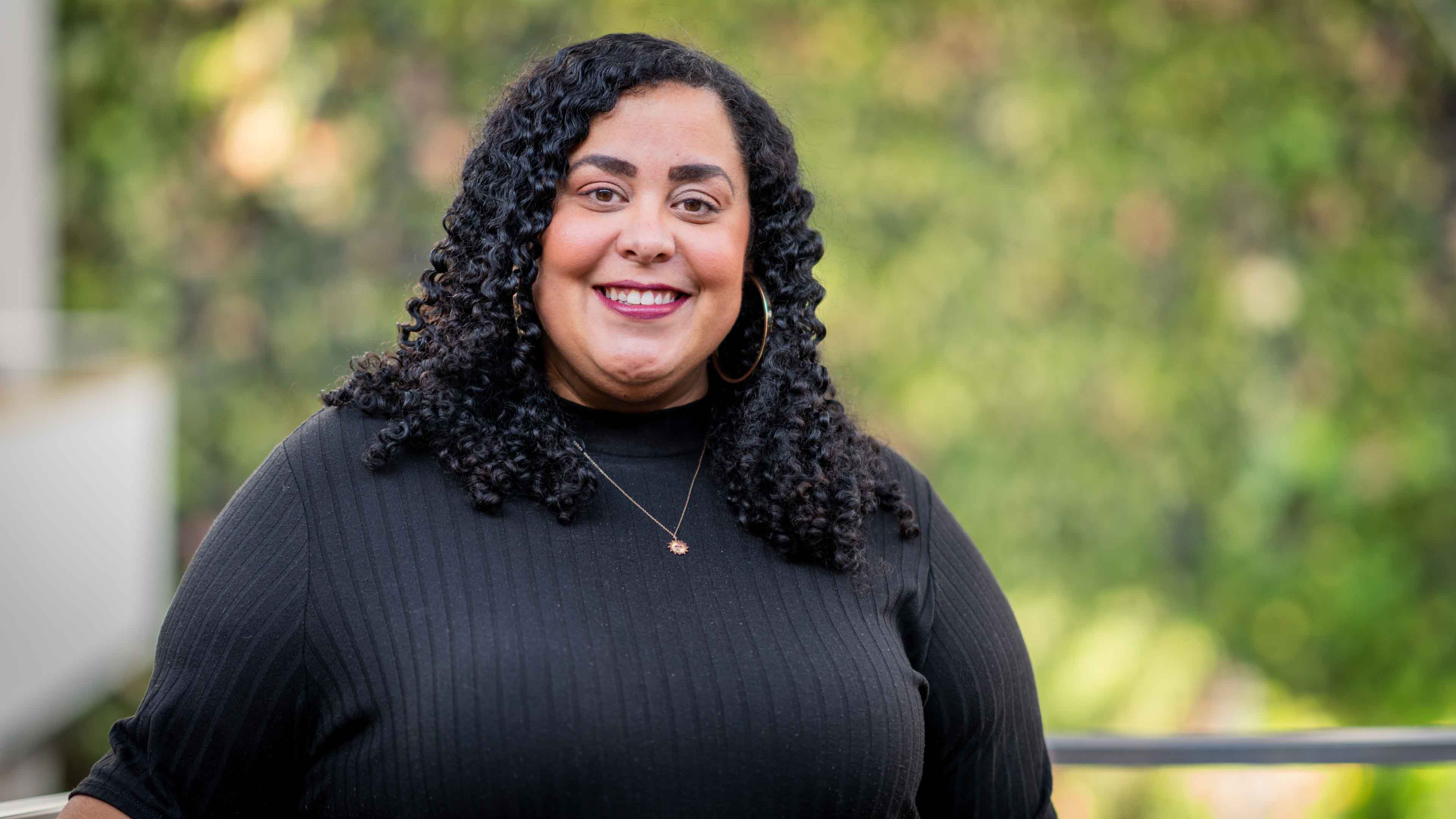 Kimberly Daniels wears a black turtleneck and smiles in front of the green plant wall