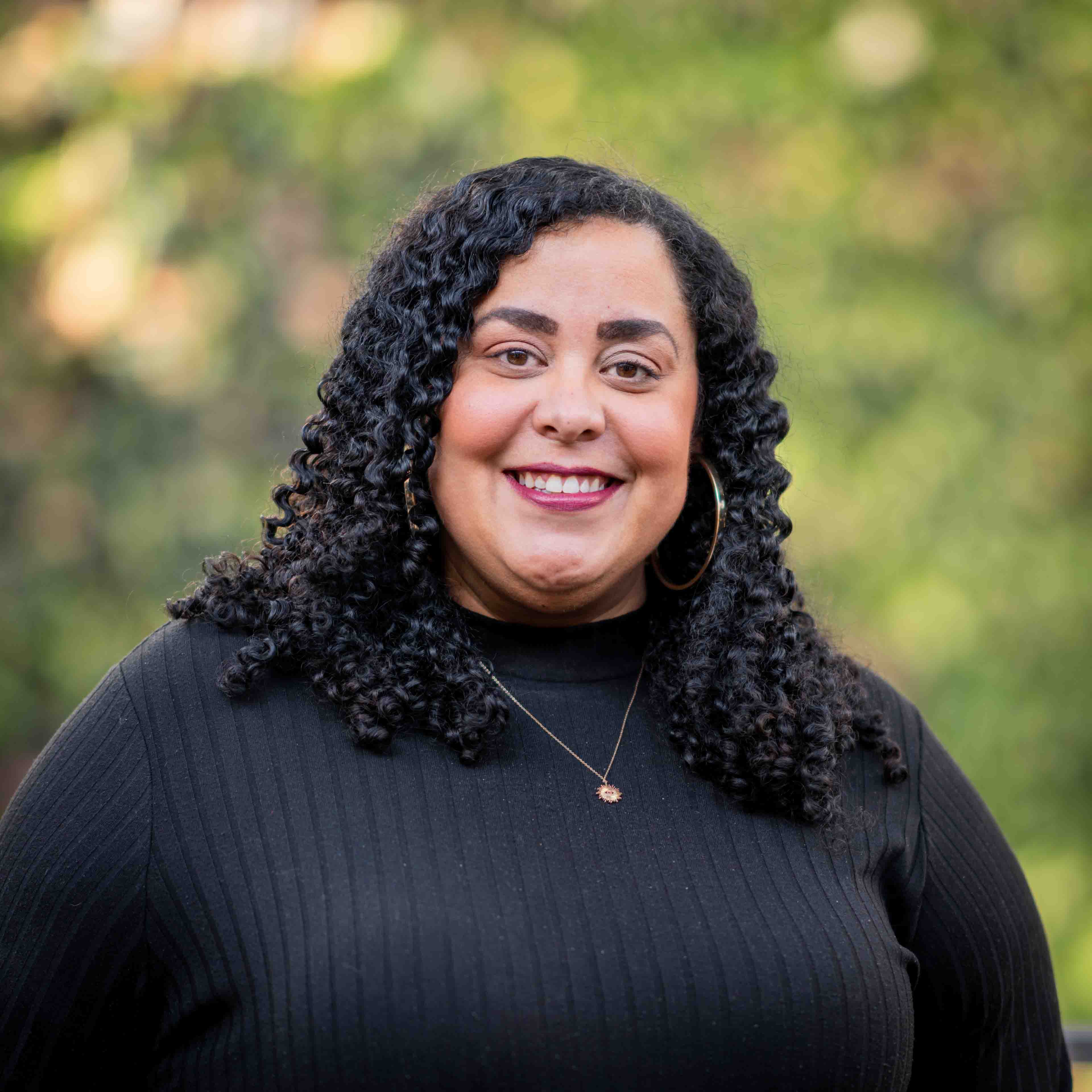Kimberly Daniels wears a black turtleneck and smiles in front of the green plant wall