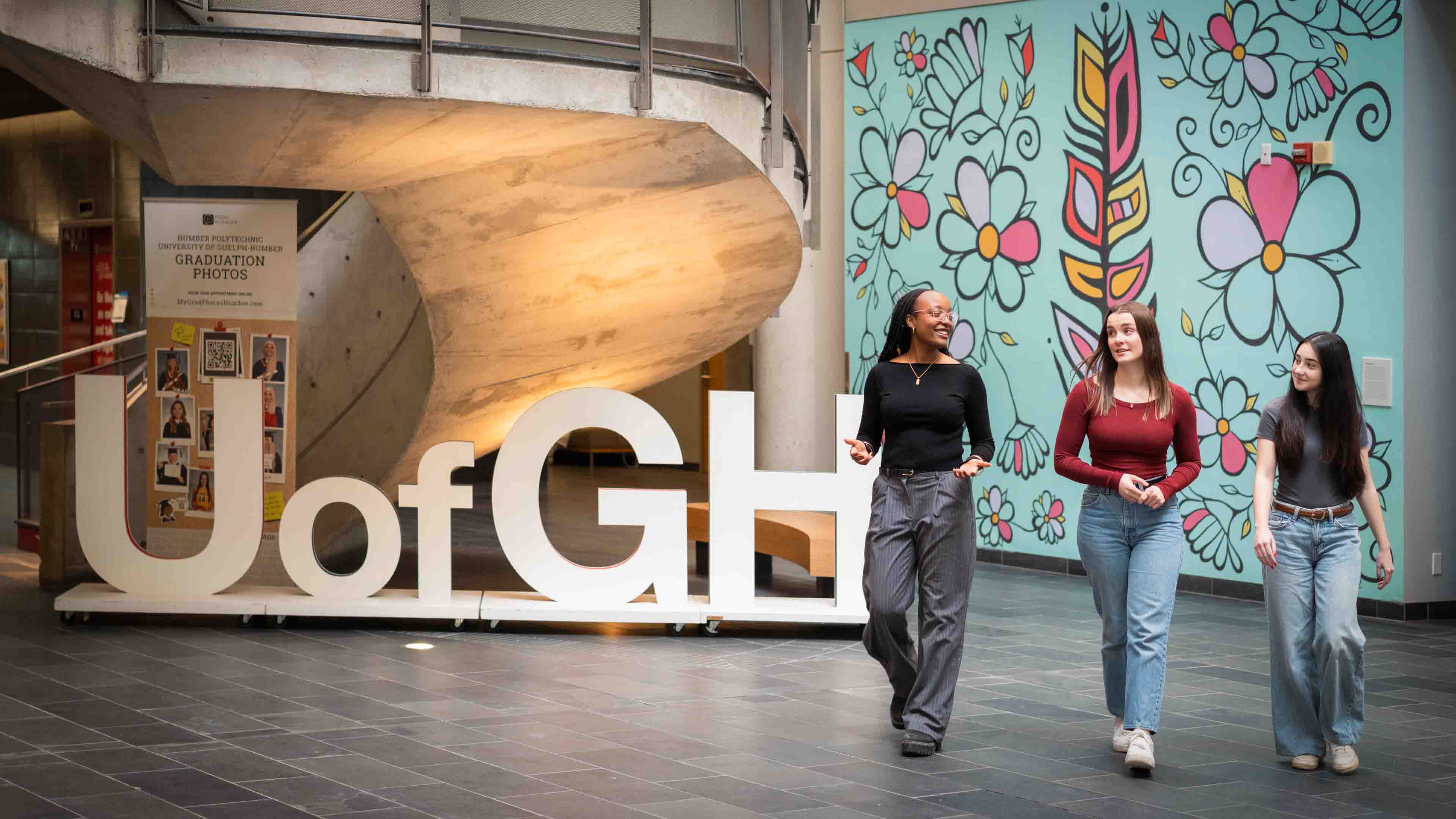 Three students walk in the atrium in front of the U of GH letter sign