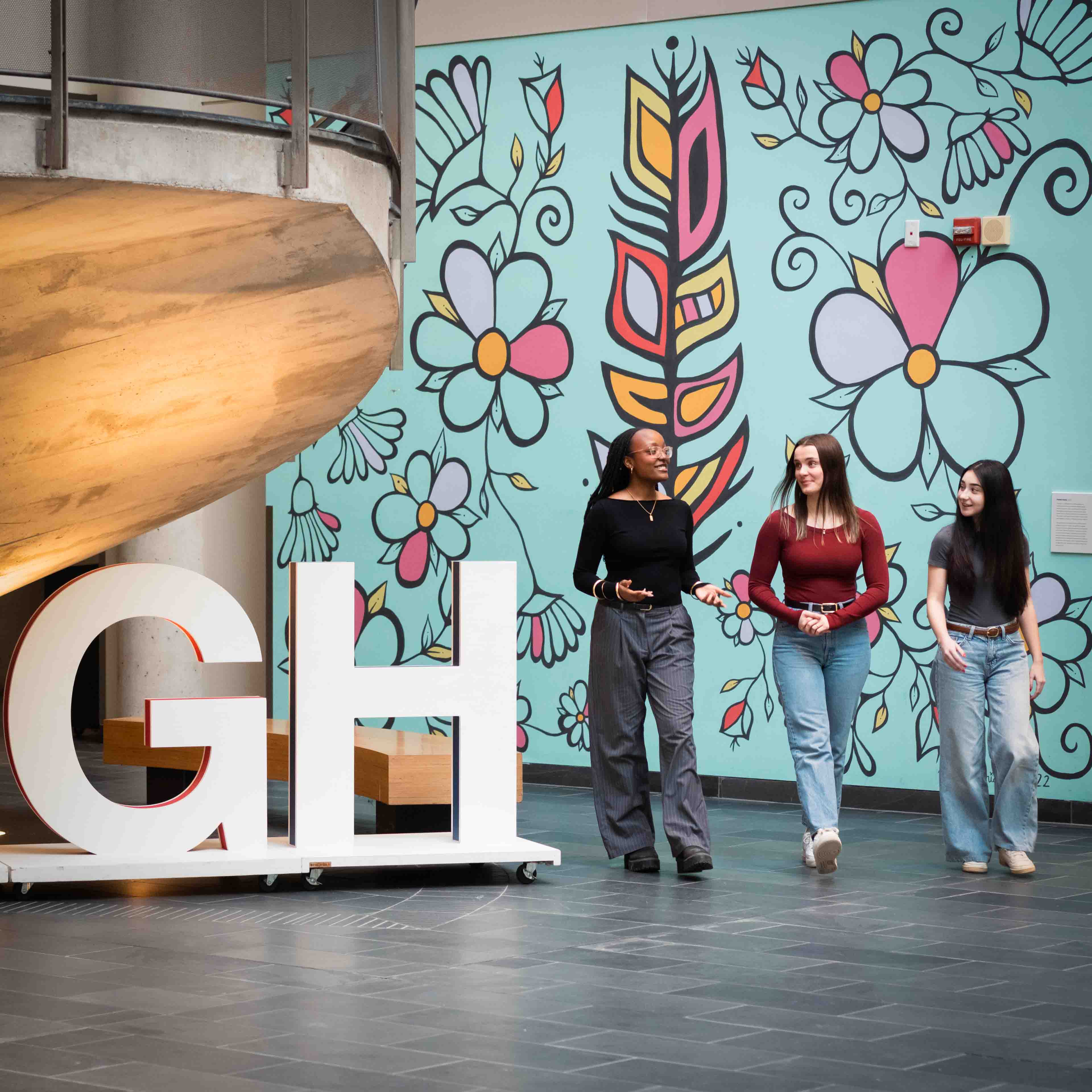Three students walk in the atrium in front of the U of GH letter sign