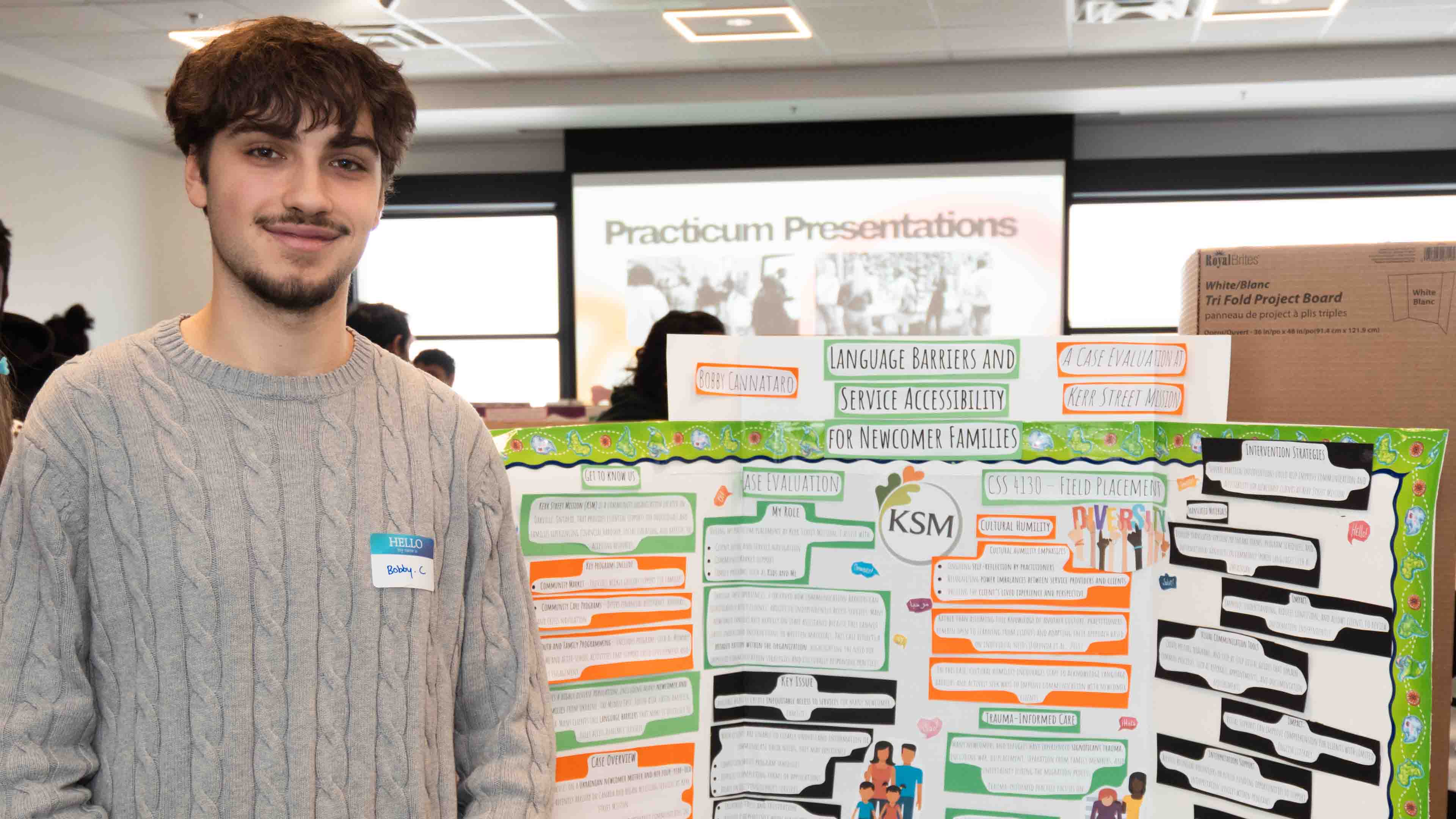 A student poses with his presentation board and a sign behind him reads "practicum presentations