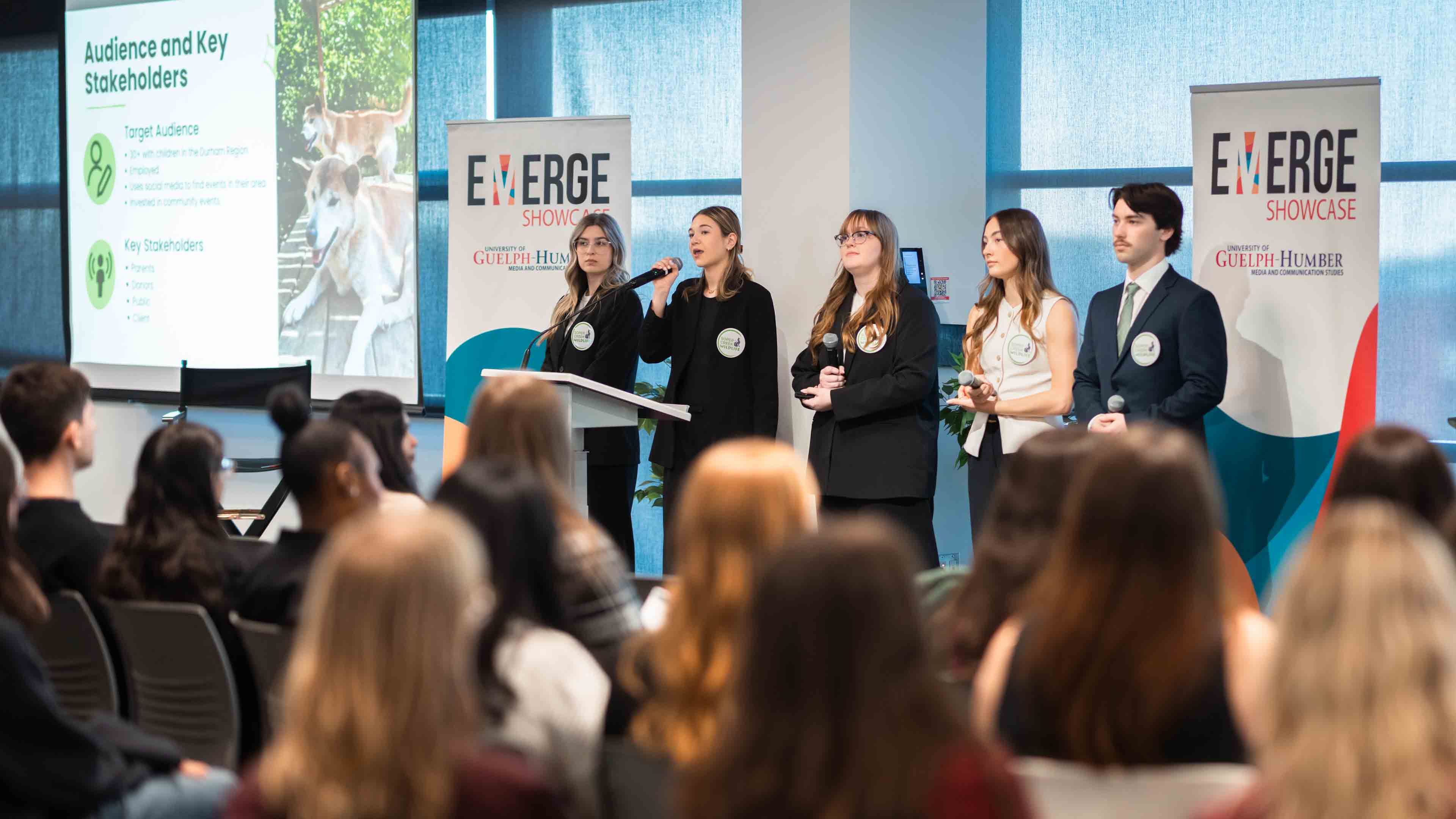 Five students stand up at the front of the room presenting at EMERGE