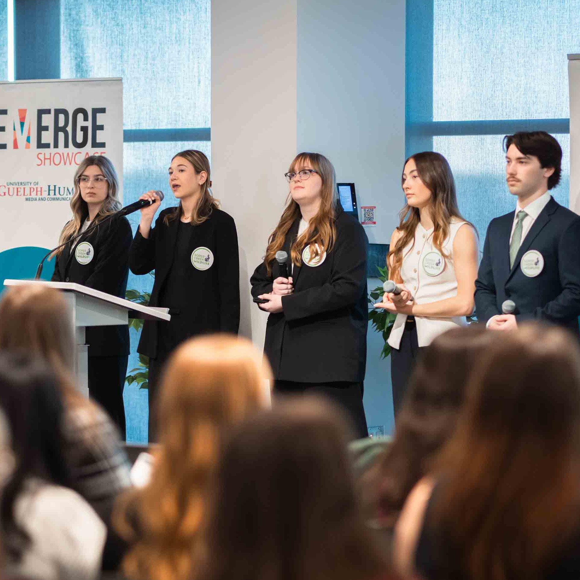 Five students stand up at the front of the room presenting at EMERGE