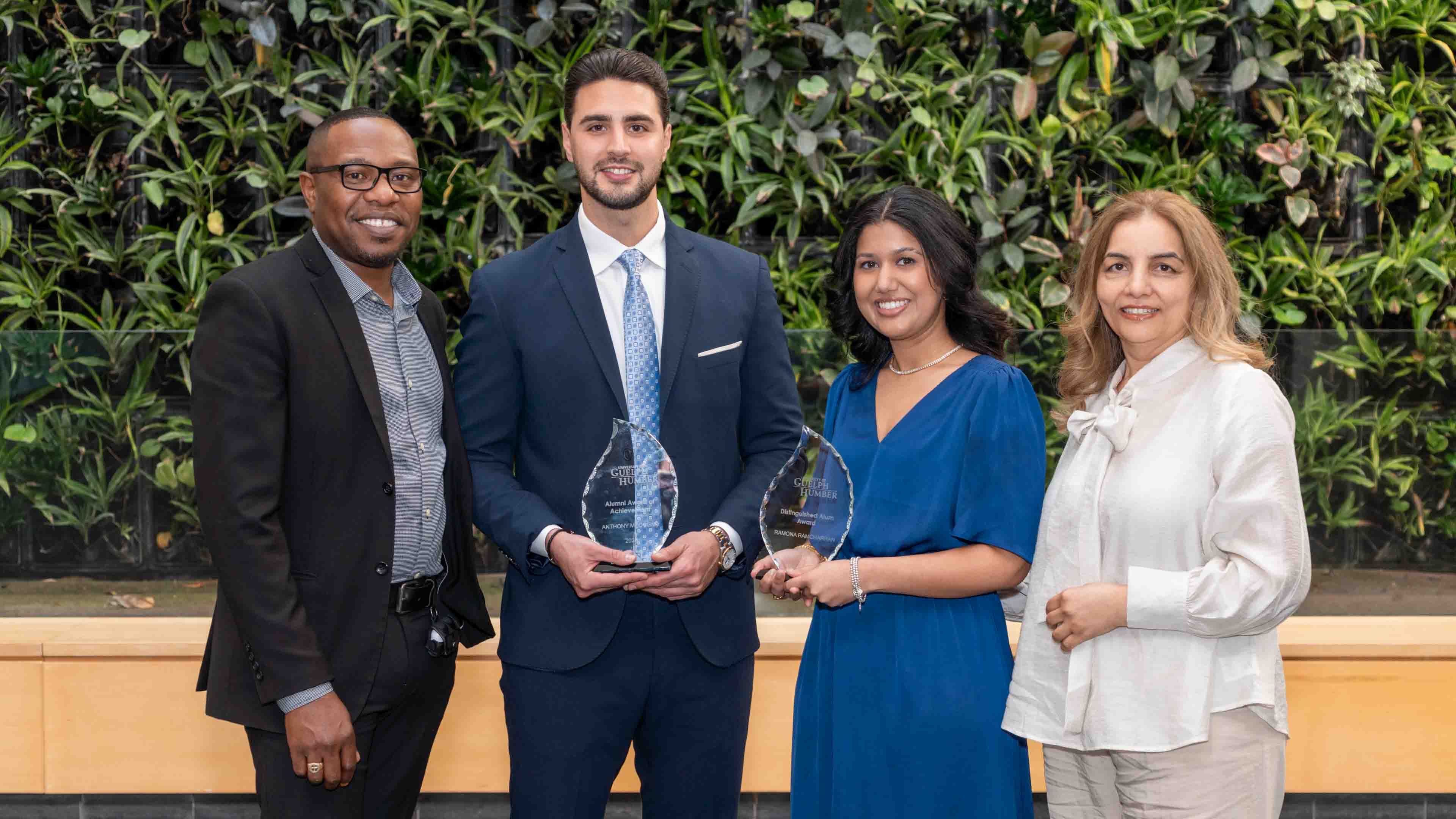 The two alumni recipients hold their awards in front of the plant wall, flanked by two members of the Business program's leadership