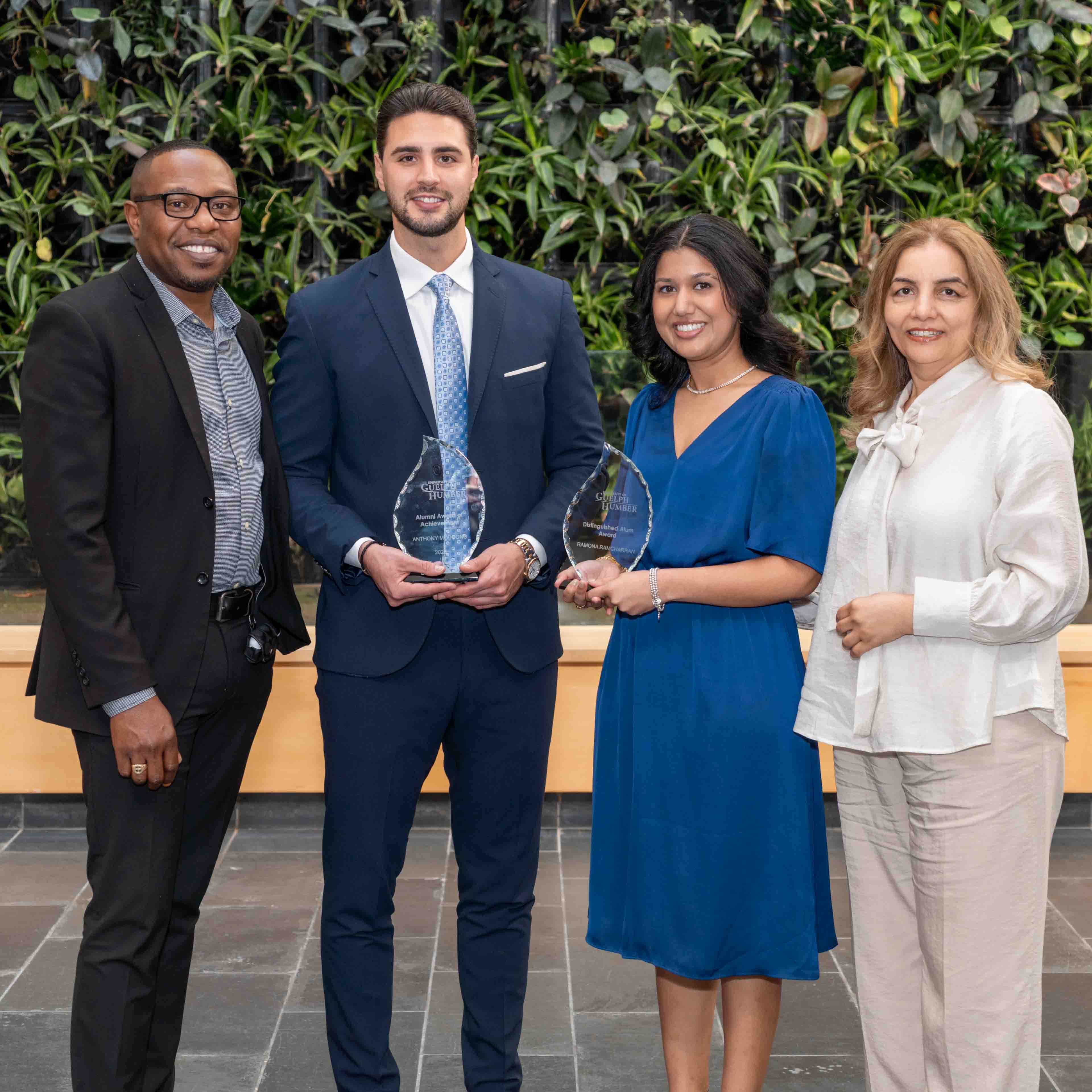 The two alumni recipients hold their awards in front of the plant wall, flanked by two members of the Business program's leadership