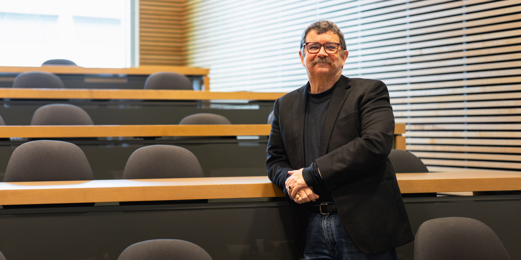 Glenn Hanna leaning against a desk in a classroom, smiling.