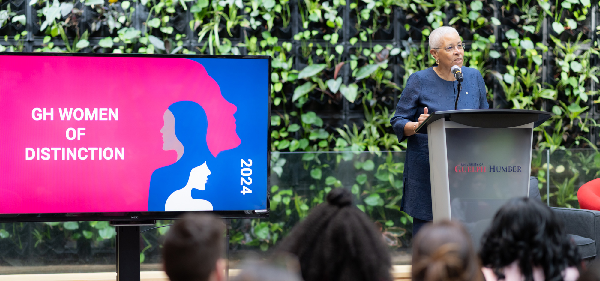 An individual standing at a podium addressing the audience with a TV on their right the reads, "GH Women of Distinction"