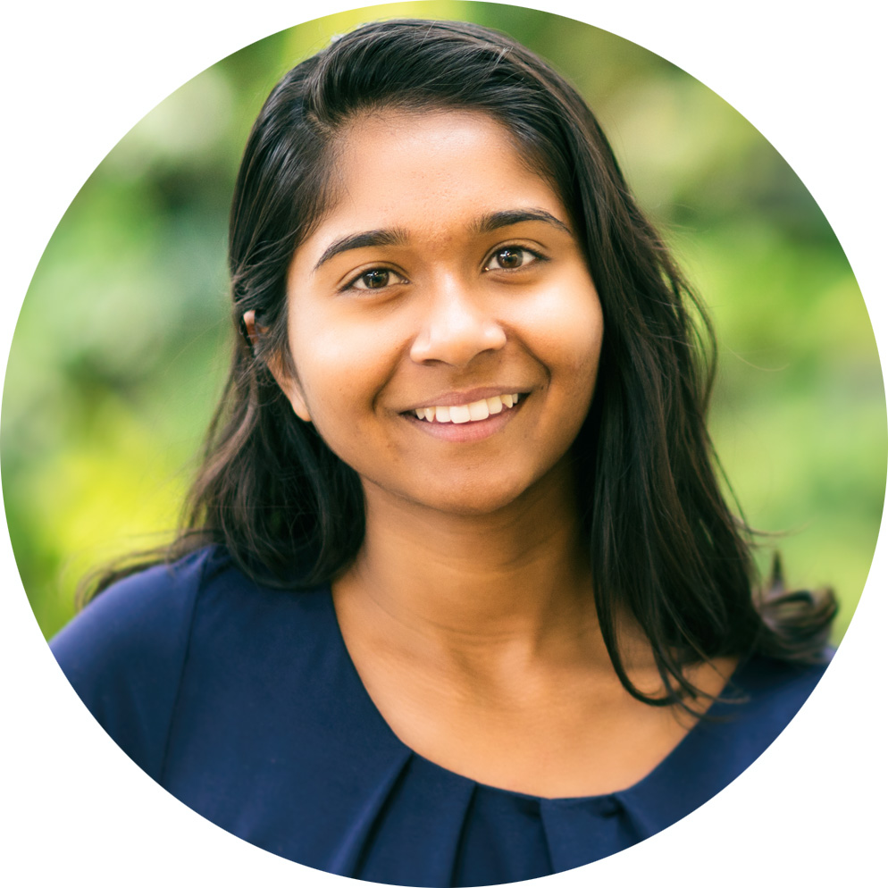 Priya Rajkumar smiles warmly by the plant wall